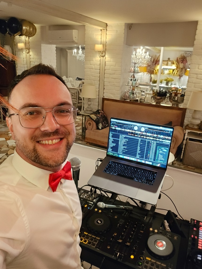 Uśmiechnięty DJ w muszce, z laptopem i sprzętem DJ-skim na tle eleganckiego wnętrza z dekoracjami. Widoczny mikrofon i elementy nagłośnienia.