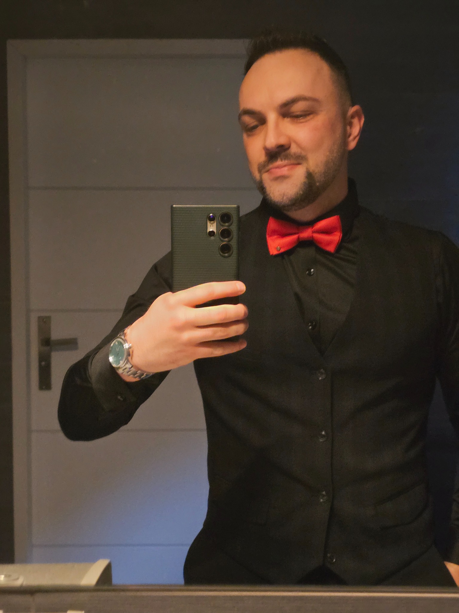 Mężczyzna w czarnej kamizelce i czerwonej muszce robi selfie telefonem. Widoczny zegarek na ręce i fragment łazienki w tle.