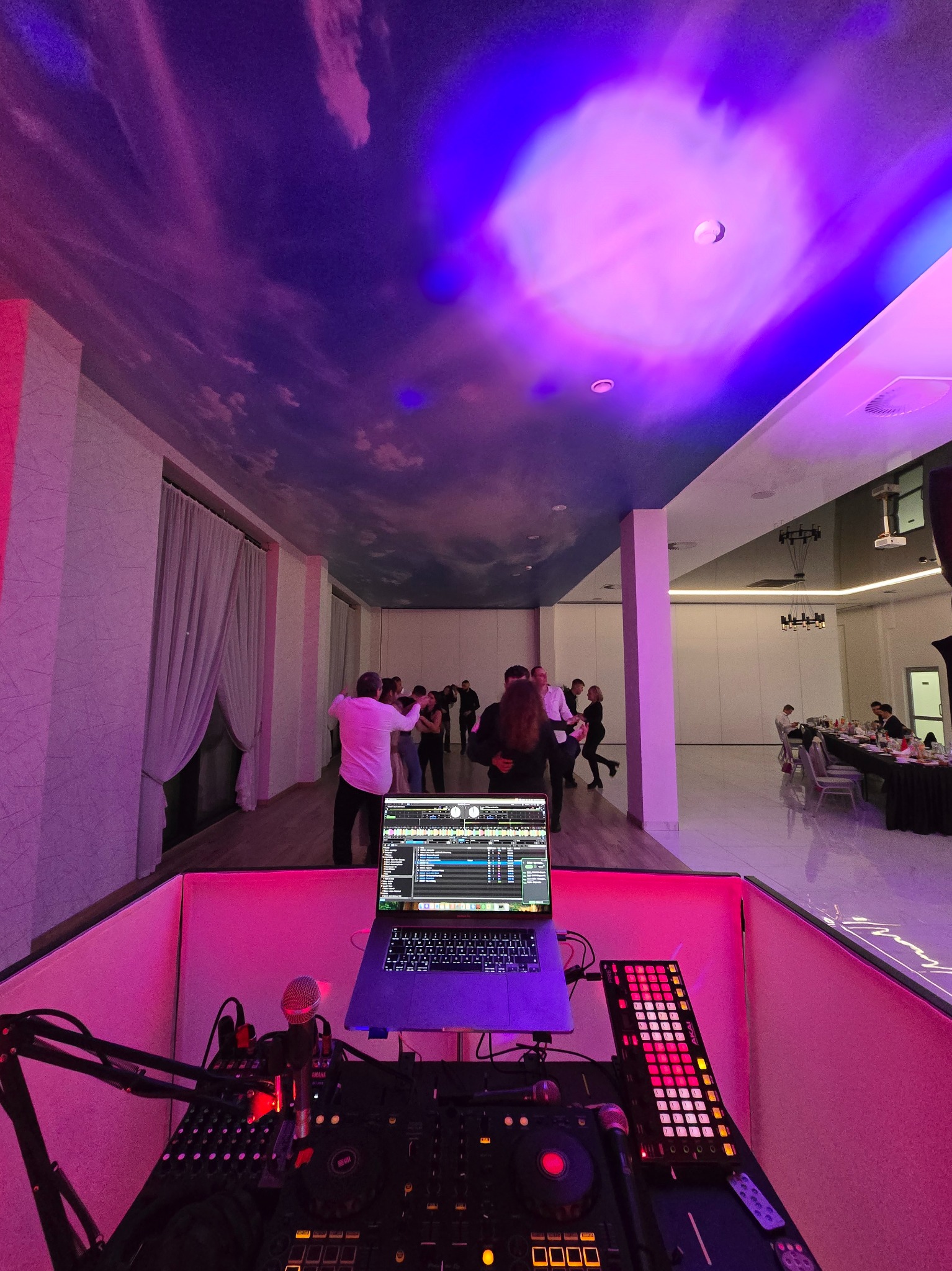 Stanowisko DJ-a z laptopem i mikserem na tle tańczących osób w sali z dekoracyjnym oświetleniem i malowanym sufitem. Widok z perspektywy DJ-a.