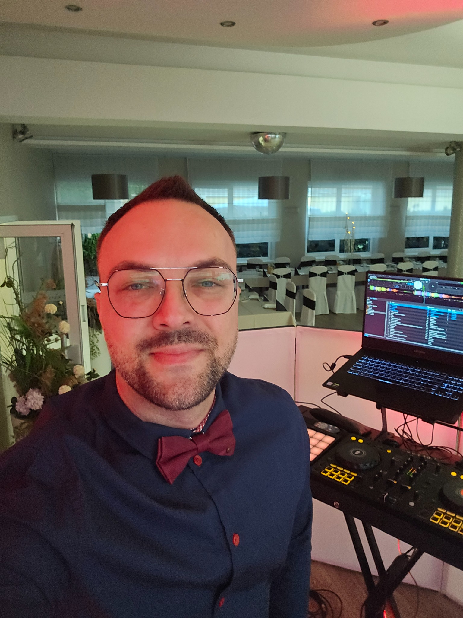 DJ z muszką, w okularach, robiący selfie na tle sali z laptopem i sprzętem DJ-skim. W tle dekoracje i parkiet. Czerwone oświetlenie sceniczne.
