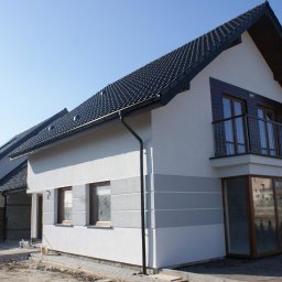 Beautyfull House - Dwukondygnacyjny dom z balkonem, wykończony w bieli i szarości, z ciemną dachówką. Widoczne elementy drewniane i metalowe balustrady. Teren wokół domu w trakcie zagospodarowania.