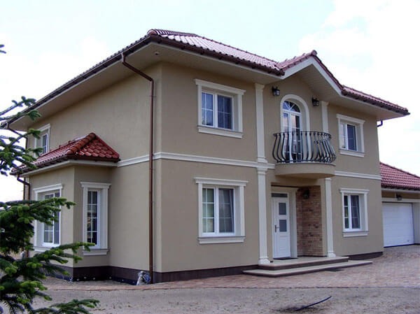 Dwukondygnacyjny dom z beżową elewacją, białymi obramowaniami okien i balkonem z ozdobną balustradą. Czerwona dachówka i brązowe rynny.