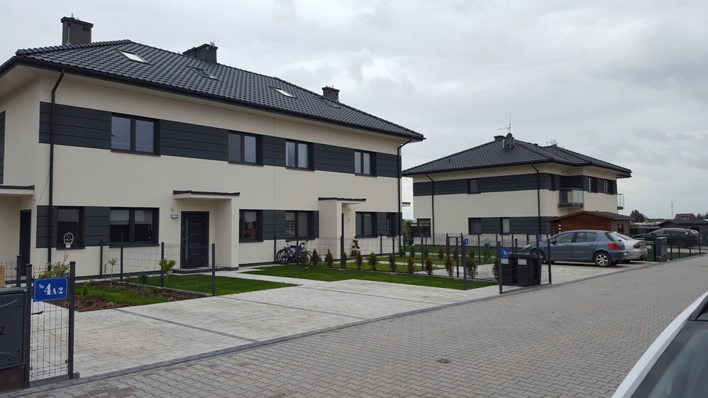 Dwa nowoczesne, dwukondygnacyjne domy bliźniacze z jasną elewacją i ciemnymi pasami, widoczne z perspektywy ulicy, z zadbanym trawnikiem i kostką brukową.