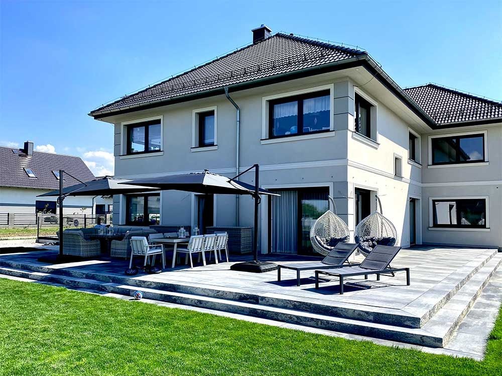 Dwukondygnacyjny dom z szarą elewacją, widok z ogrodu. Na tarasie meble ogrodowe, huśtawki kokony i leżaki. Trawa, błękitne niebo. Nowoczesny styl.