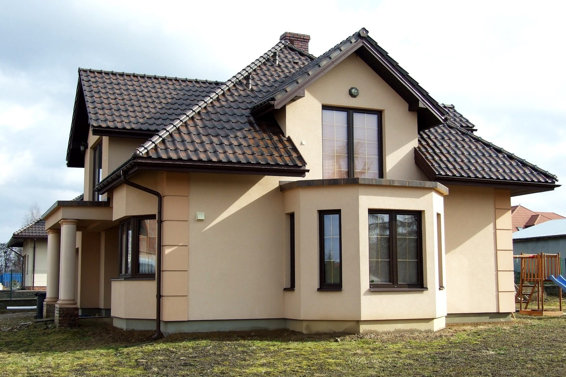 Dwukondygnacyjny dom z beżową elewacją i ciemnobrązowym dachem. Widoczne okna z ciemnymi ramami i kolumny przy wejściu. Na trawniku widać resztki śniegu.