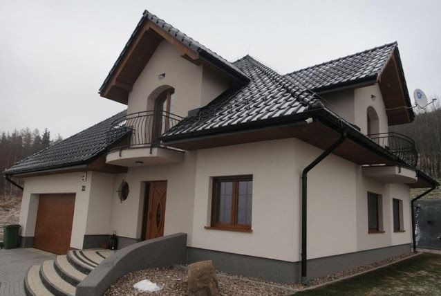 Dwupiętrowy dom z beżową elewacją, czarnym dachem i balkonami z kutymi balustradami. Widoczne schody wejściowe i garaż. Tło stanowią drzewa i pochmurne niebo.