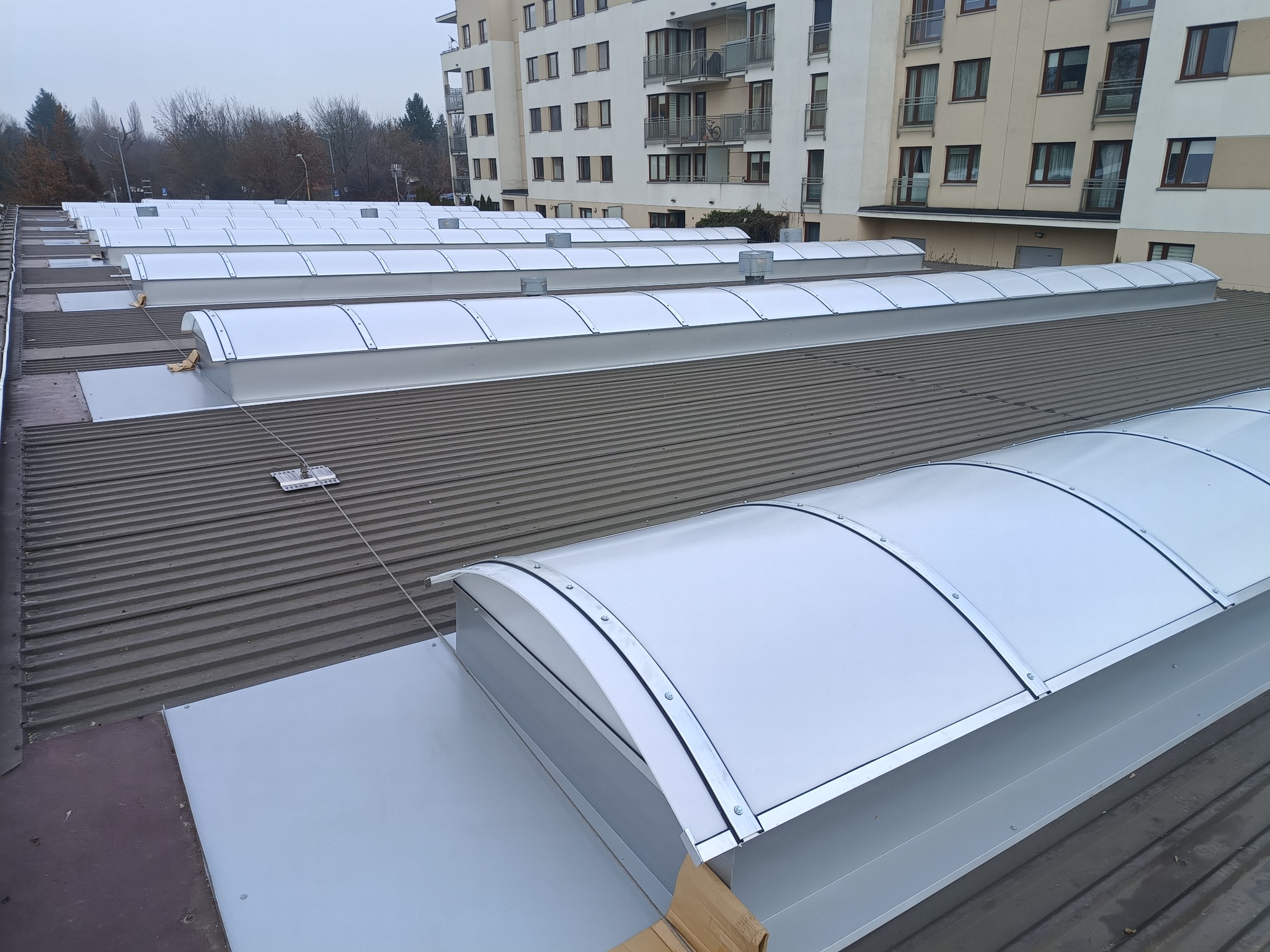 Szary dach budynku z zamontowanymi świetlikami dachowymi o półokrągłym kształcie. W tle widoczny budynek mieszkalny i drzewa.