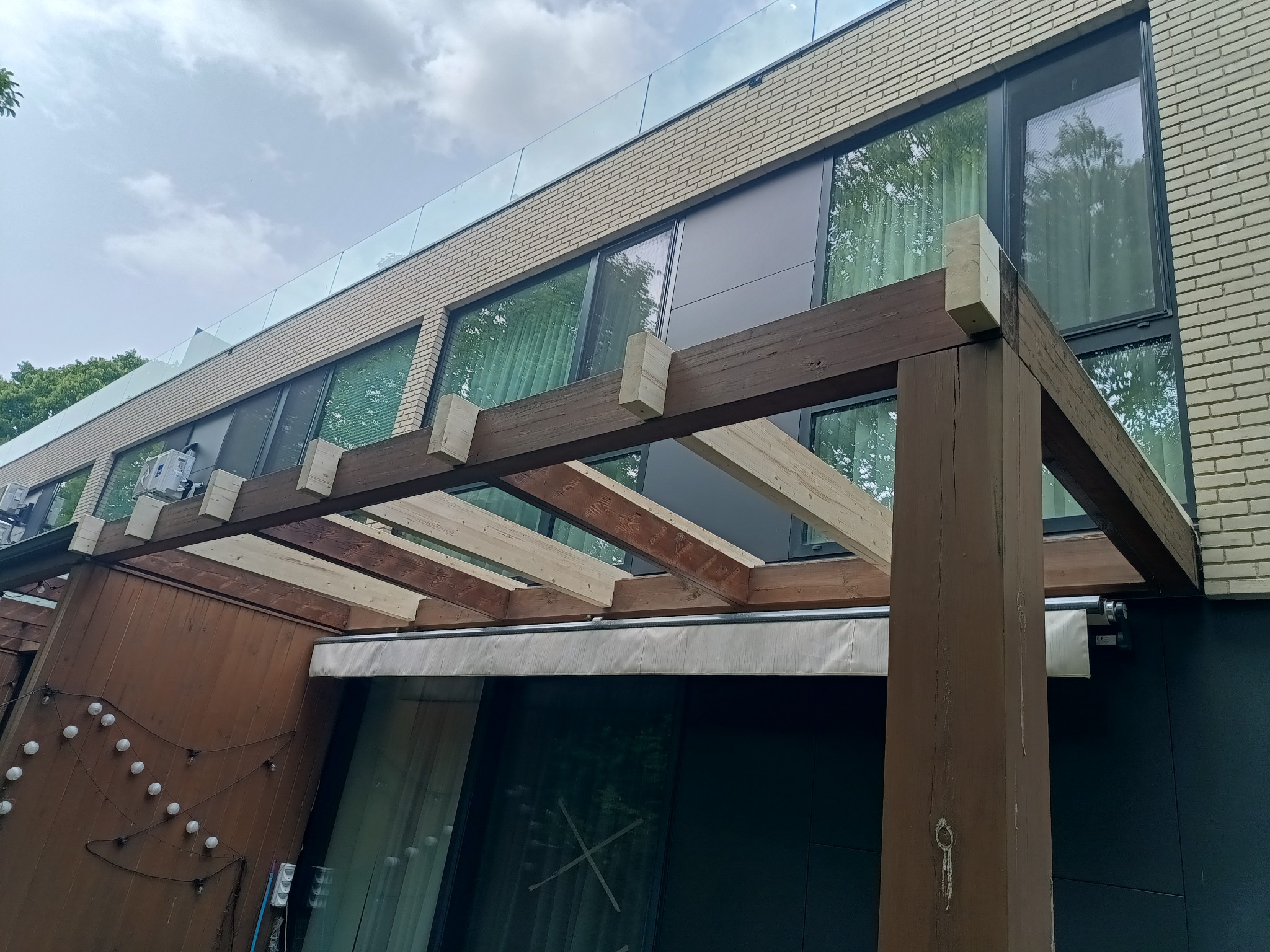 Nowoczesna altana z drewnianą konstrukcją, przylegająca do budynku z dużymi oknami i szklaną balustradą. Widoczne elementy oświetlenia i markiza.