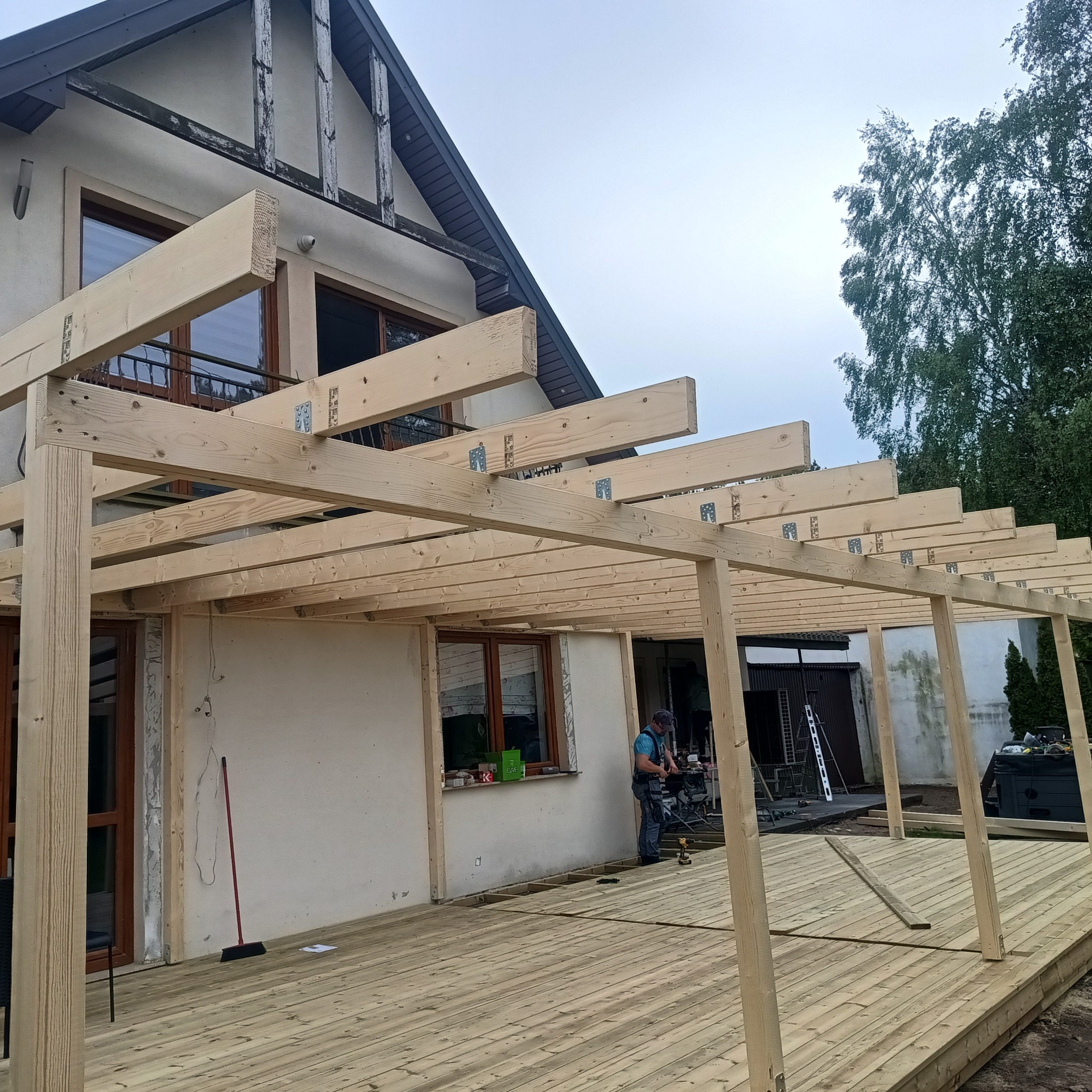 Drewniana konstrukcja altany w trakcie budowy, widoczna z zewnątrz. Na pierwszym planie deski podłogowe, w tle dom jednorodzinny i pracownik w trakcie montażu.
