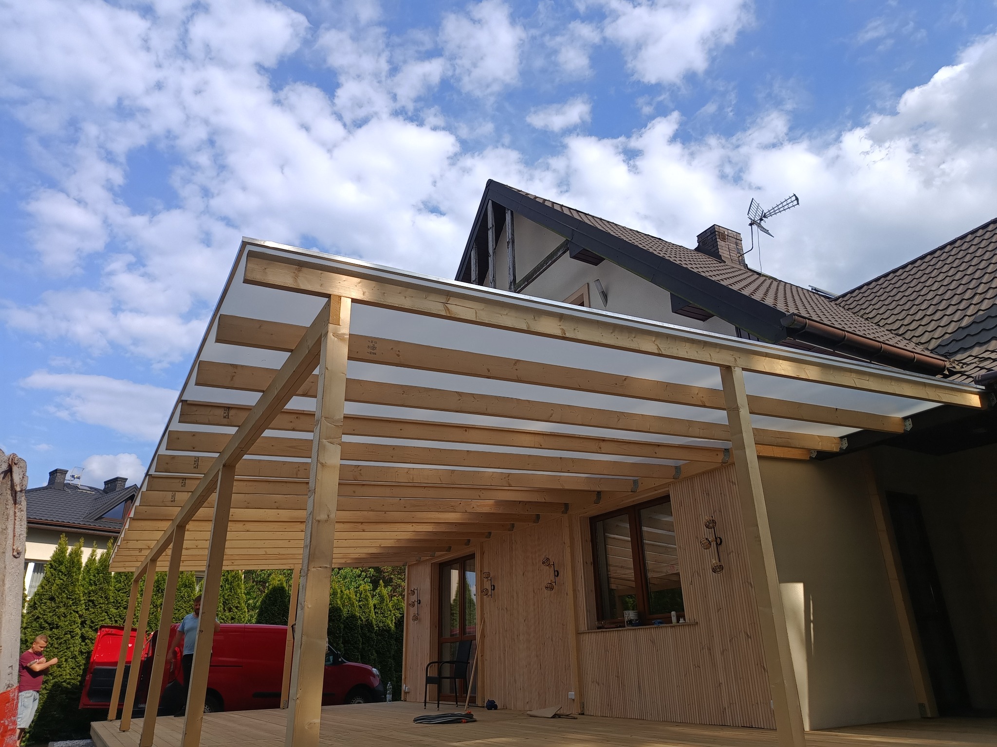 Nowoczesna altana z drewnianą konstrukcją i przezroczystym zadaszeniem, widoczna na tle błękitnego nieba z chmurami. Czerwony samochód dostawczy w tle sugeruje etap budowy.
