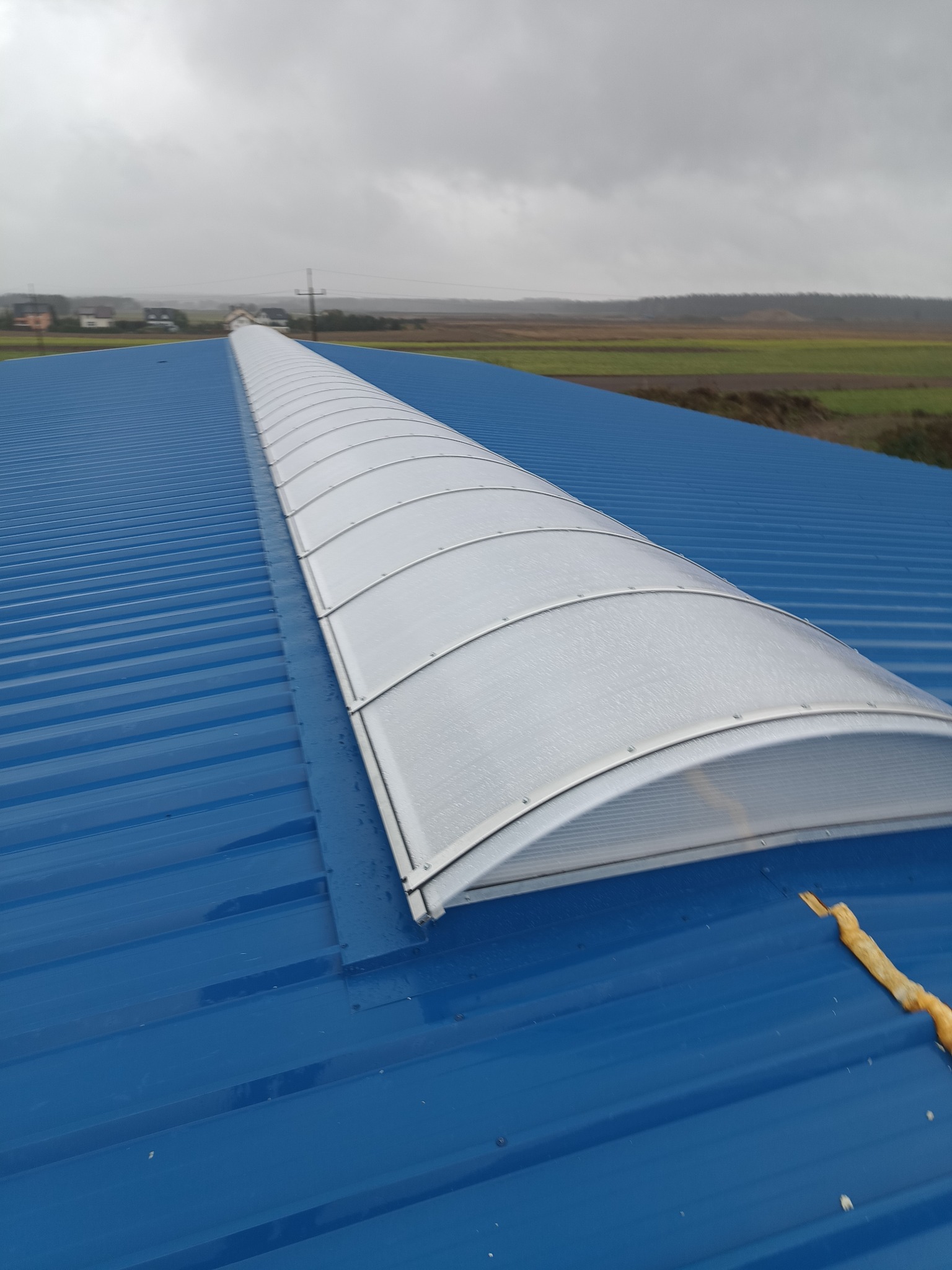 Niebieski dach z blachy trapezowej z długim, łukowym świetlikiem z tworzywa sztucznego. W tle widoczne pola i zabudowania w pochmurny dzień.