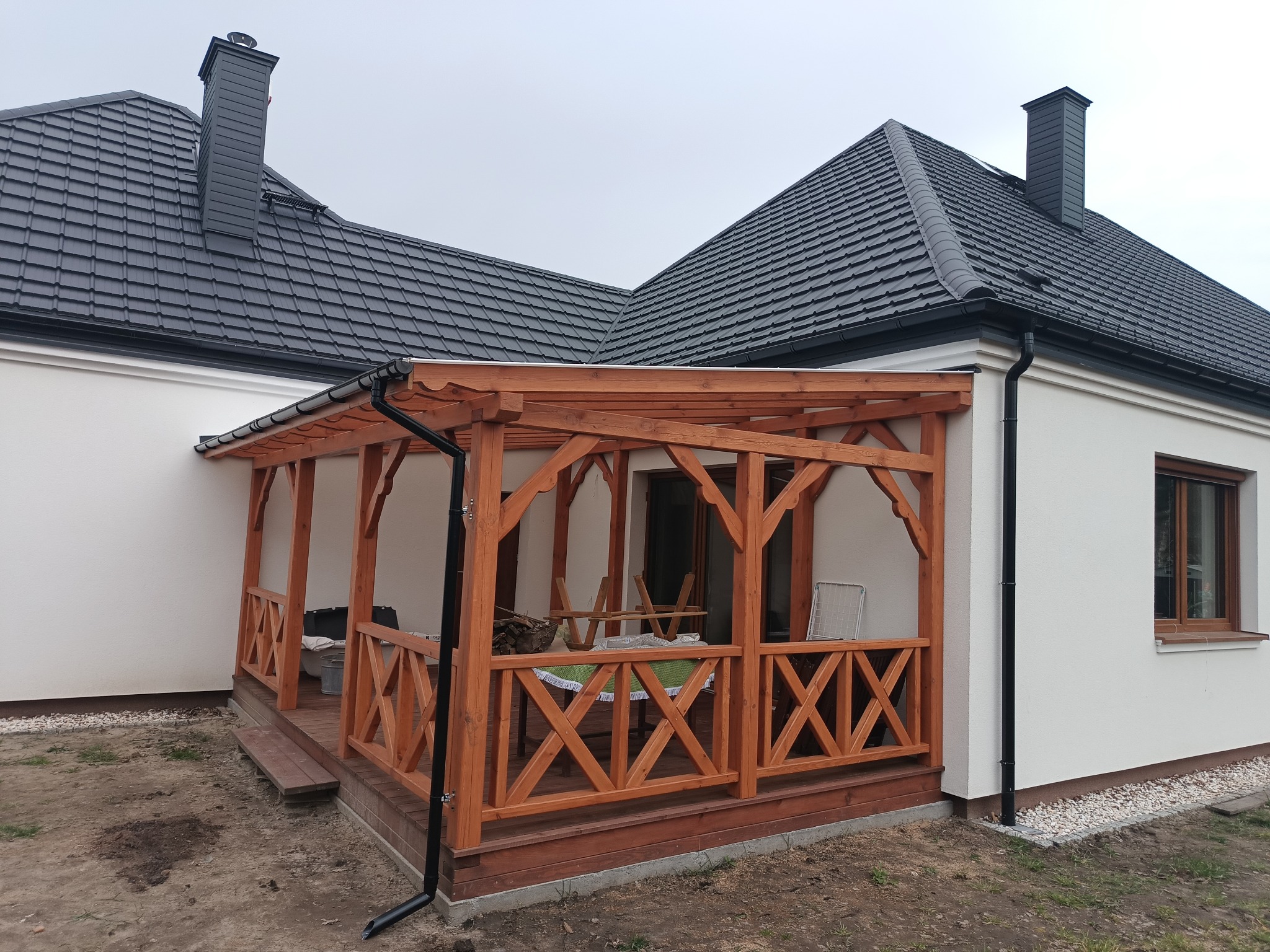 Drewniana altana z balustradą w kształcie litery X, przylegająca do białego domu z grafitowym dachem. Widoczny fragment rynny i okna. Uwagę przykuwa staranne wykonanie.