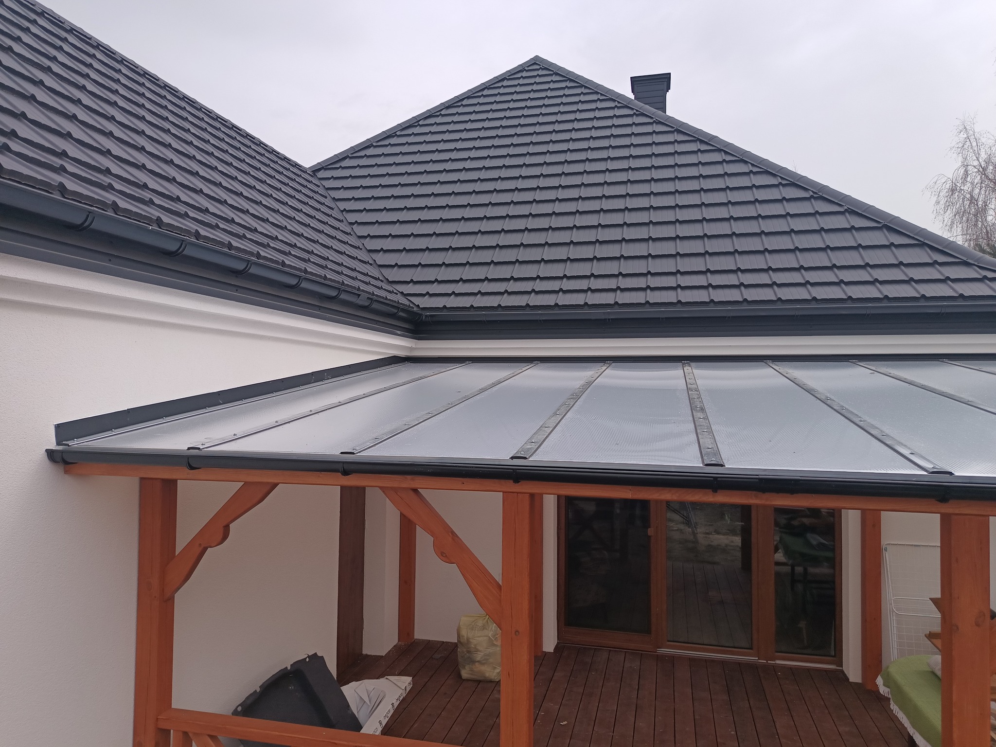 Nowoczesna altana z drewnianą konstrukcją i przezroczystym zadaszeniem, przylegająca do budynku z ciemnoszarym dachem z dachówki ceramicznej.