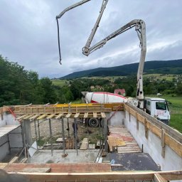 M.K.W HOME - Wysoki kąt: Fundamenty domu z szalunkiem i zbrojeniem, betonowóz w tle, krajobraz pagórkowaty. Widoczne podpory szalunku i częściowo zalany beton.