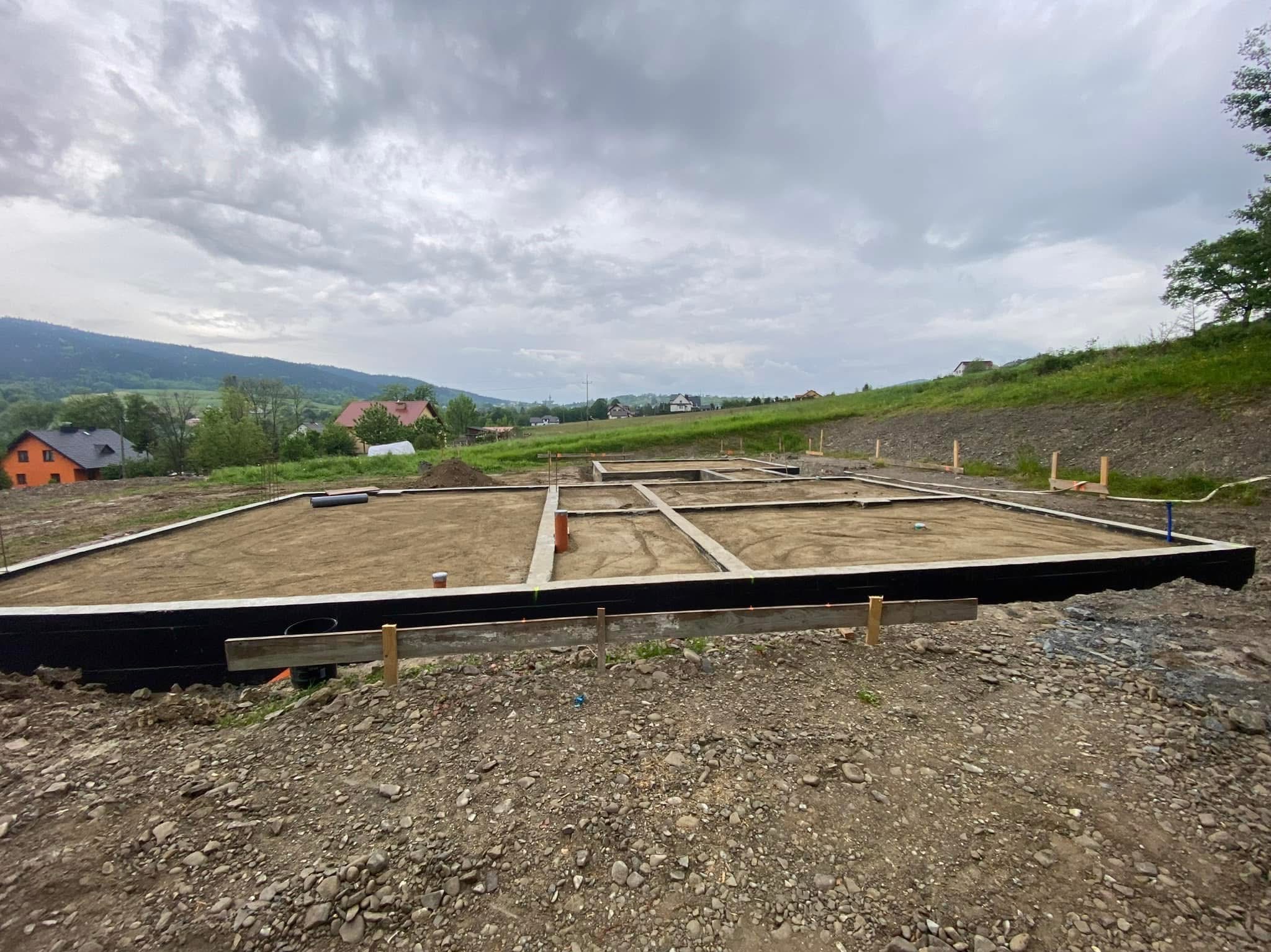 Przygotowany teren pod fundamenty domu z widoczną izolacją przeciwwilgociową i zarysowanymi ścianami działowymi na tle pagórkowatego krajobrazu.