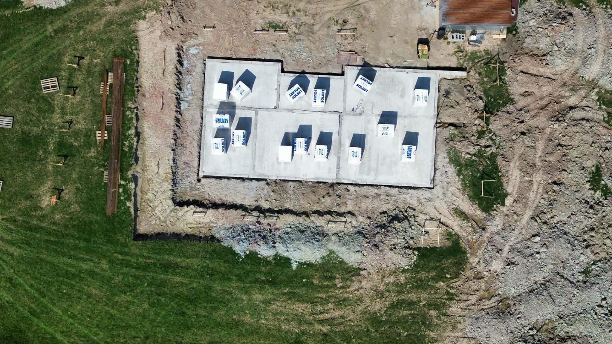 Widok z góry na wylany fundament z bloczkami Solbet, otoczony wykopem i zielenią. Palety i drewno leżą na trawie. Świeży beton.