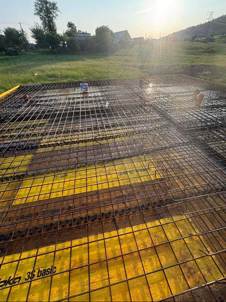 Zbrojenie płyty fundamentowej na żółtym szalunku, widok z góry. W tle zielone wzgórza i błękitne niebo z promieniami słońca. Prace budowlane w toku.