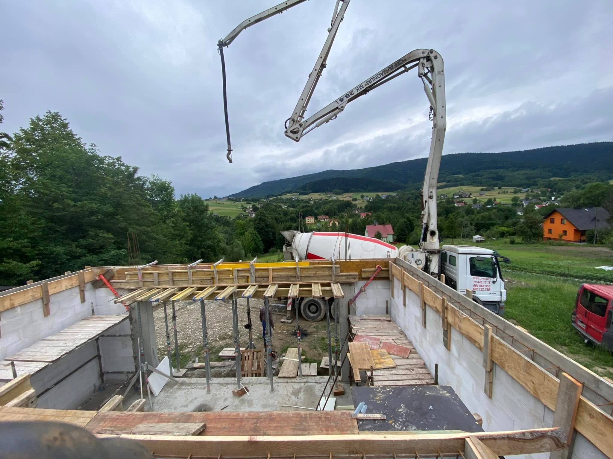 Wysoki kąt: Fundamenty domu z szalunkiem i zbrojeniem, betonowóz w tle, krajobraz pagórkowaty. Widoczne podpory szalunku i częściowo zalany beton.