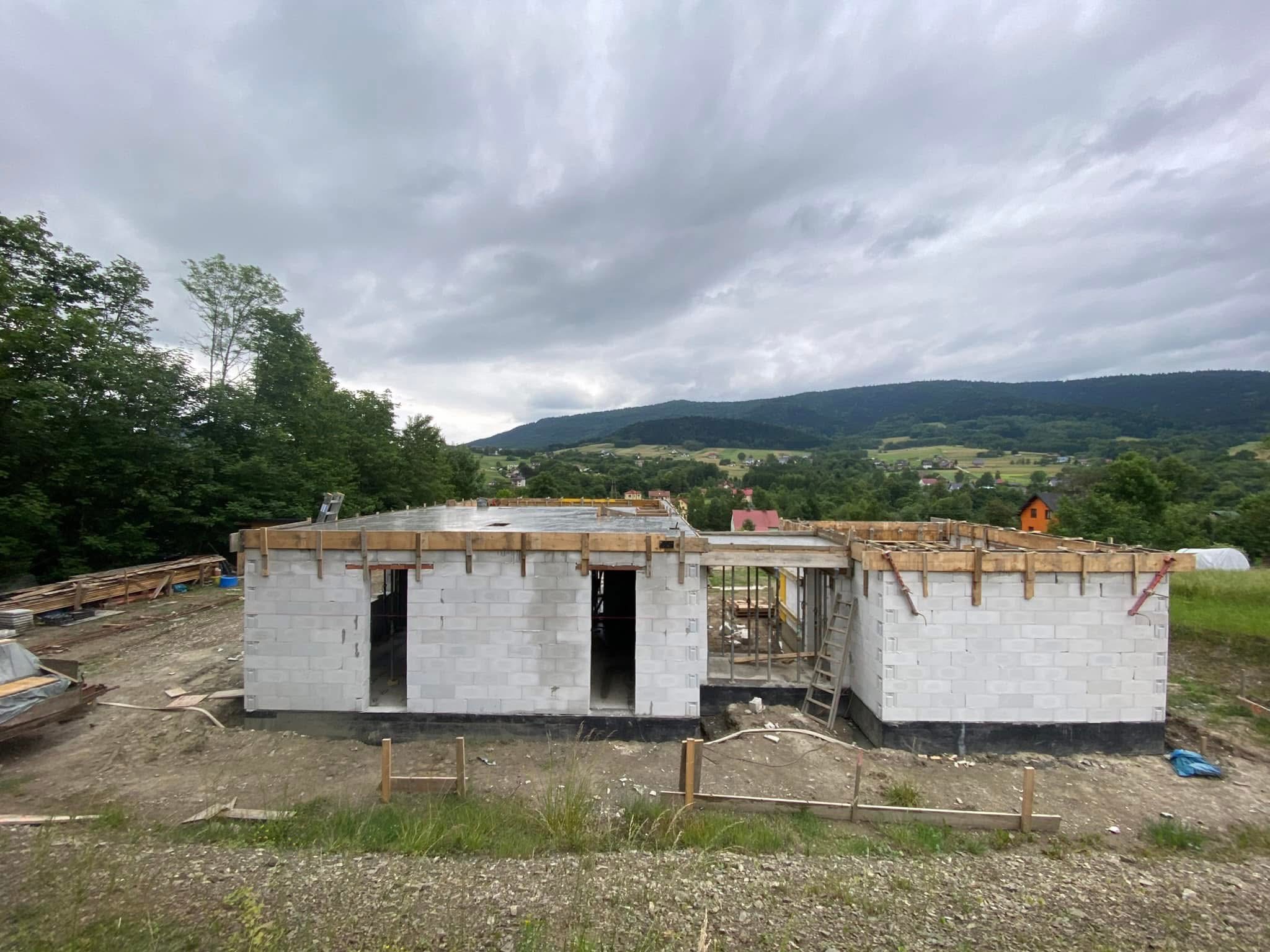 Budowa domu jednorodzinnego z białych bloczków, widoczne zbrojenia i drewniane szalunki, w tle zielone wzgórza i pochmurne niebo. Etap murowania ścian.