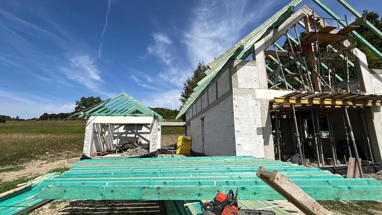 Budowa domu w stanie surowym otwartym: ściany z betonu komórkowego, drewniana konstrukcja dachu, szalunki, rusztowania. W tle łąka i błękitne niebo.
