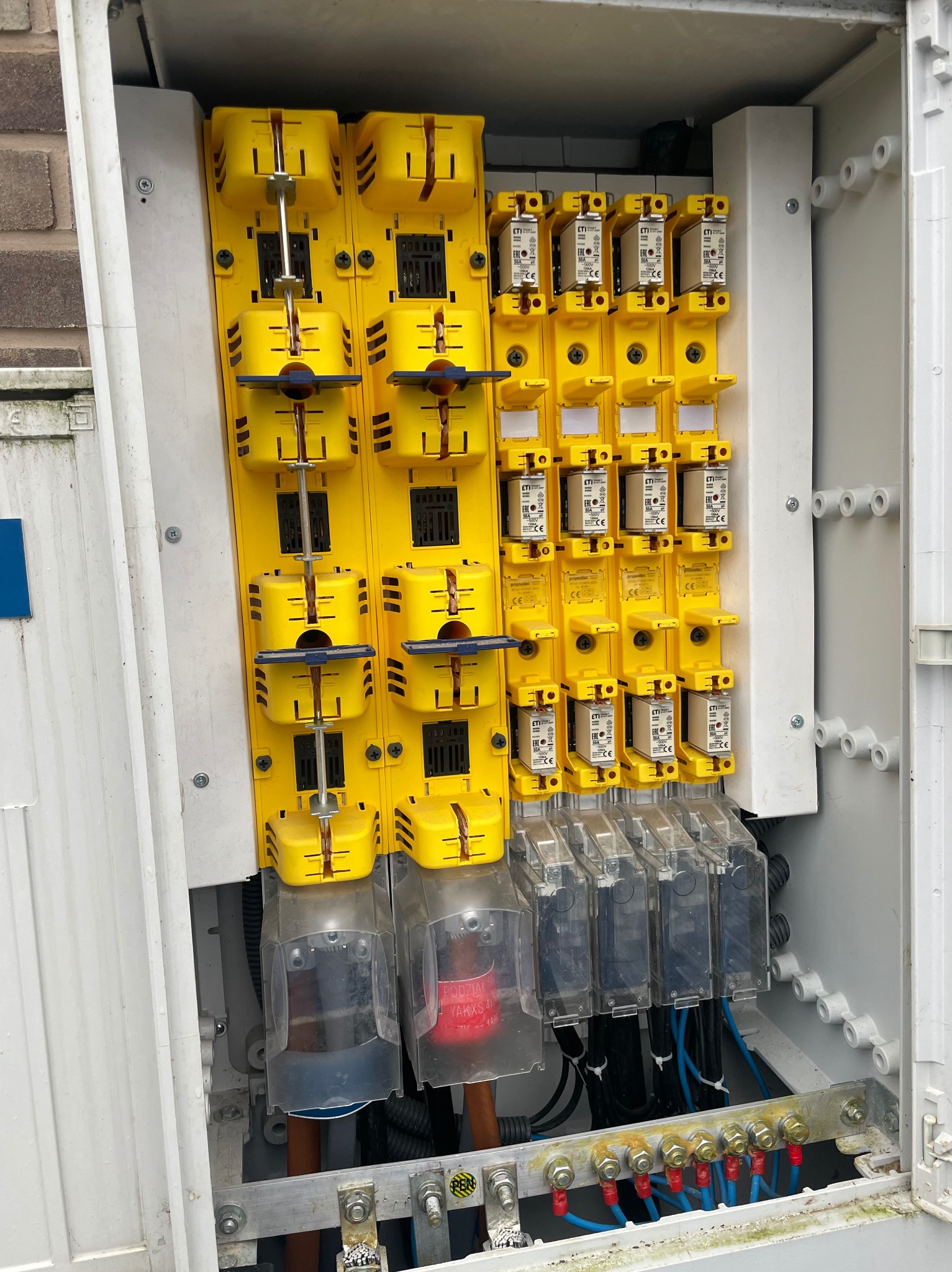 Otwarta skrzynka z żółtymi bezpiecznikami i przewodami, widoczne elementy instalacji elektrycznej. Skrzynka umieszczona na zewnątrz budynku.