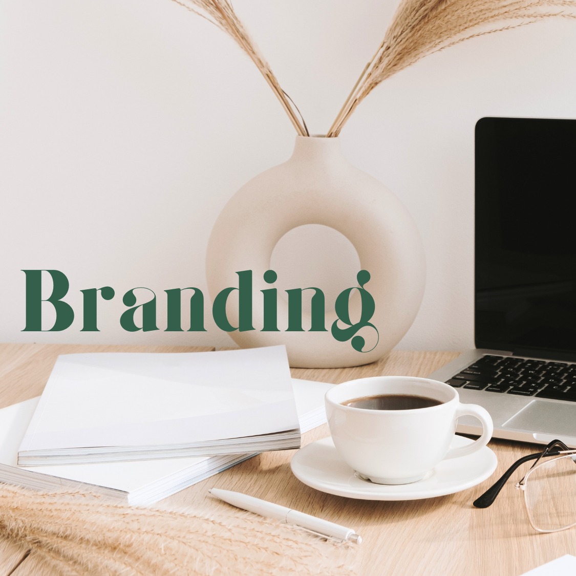 Biurko z laptopem, kawą i wazonem z trawą pampasową. Na pierwszym planie napis 'Branding' w kolorze zielonym. Stonowana kolorystyka, minimalistyczny styl.