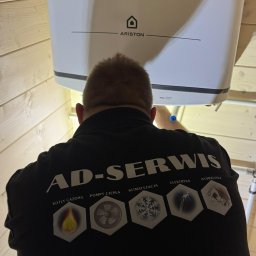 AD-SERWIS ALEKSANDRA BIELIŃSKA - Instalator w czarnej koszulce AD-SERWIS montuje biały piec gazowy Ariston w pomieszczeniu wykończonym boazerią. Widoczne rury instalacyjne.