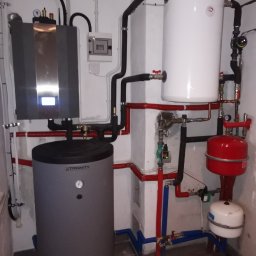 AD-SERWIS ALEKSANDRA BIELIŃSKA - Zestaw kotła gazowego z zasobnikiem ciepłej wody w kotłowni, z widocznymi rurami instalacji grzewczej i elementami zabezpieczającymi.
