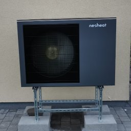 AD-SERWIS ALEKSANDRA BIELIŃSKA - Zewnętrzna jednostka pompy ciepła marki Nesheat, zamontowana na metalowej konstrukcji, na tle elewacji budynku. Widoczny fragment trawnika i kostki brukowej.