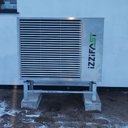 AD-SERWIS ALEKSANDRA BIELIŃSKA - Pompa ciepła IZZIFAST zamontowana na zewnątrz budynku, na betonowych podstawach, z widocznym logo firmy. Śnieg na ziemi.