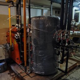 AD-SERWIS ALEKSANDRA BIELIŃSKA - Pomarańczowy piec grzewczy z czarnym zbiornikiem owiniętym folią, połączone rurami w piwnicy z szarą podłogą i białymi ścianami. Widoczne liczne zawory i manometry.