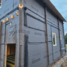 AD-SERWIS ALEKSANDRA BIELIŃSKA - Dom w trakcie budowy z widoczną instalacją elektryczną na zewnątrz. Czarne rury prowadzone po ścianach pokrytych membraną. Widoczne okna i fragment konstrukcji dachu.