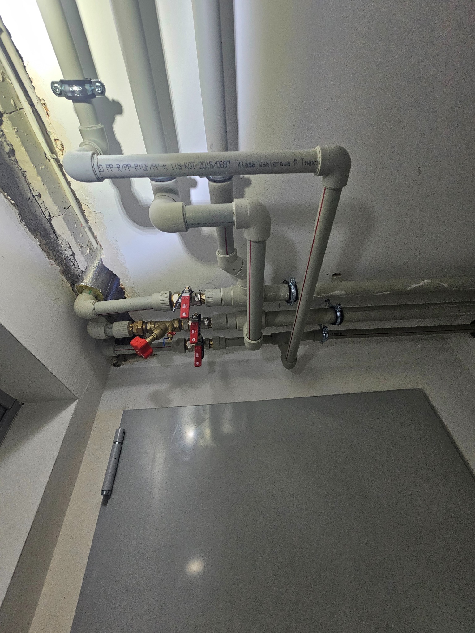 Instalacja rur z zaworami w Elblągu, widok na sufit i fragment ściany. Rury z tworzywa sztucznego, niektóre z czerwonym paskiem, zamocowane do sufitu. Widoczne zawory.