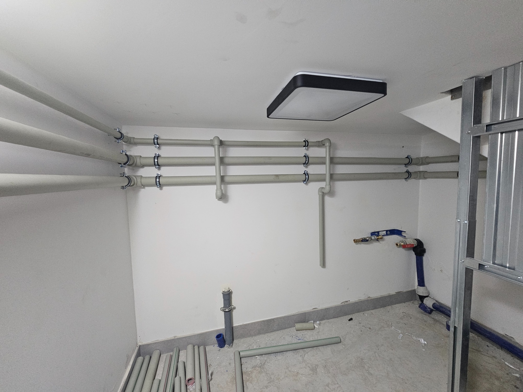 Szare rury instalacji wodnej zamocowane do białej ściany, widoczne zawory i fragmenty konstrukcji metalowej. Część rur leży na podłodze. Oświetlenie sufitowe.
