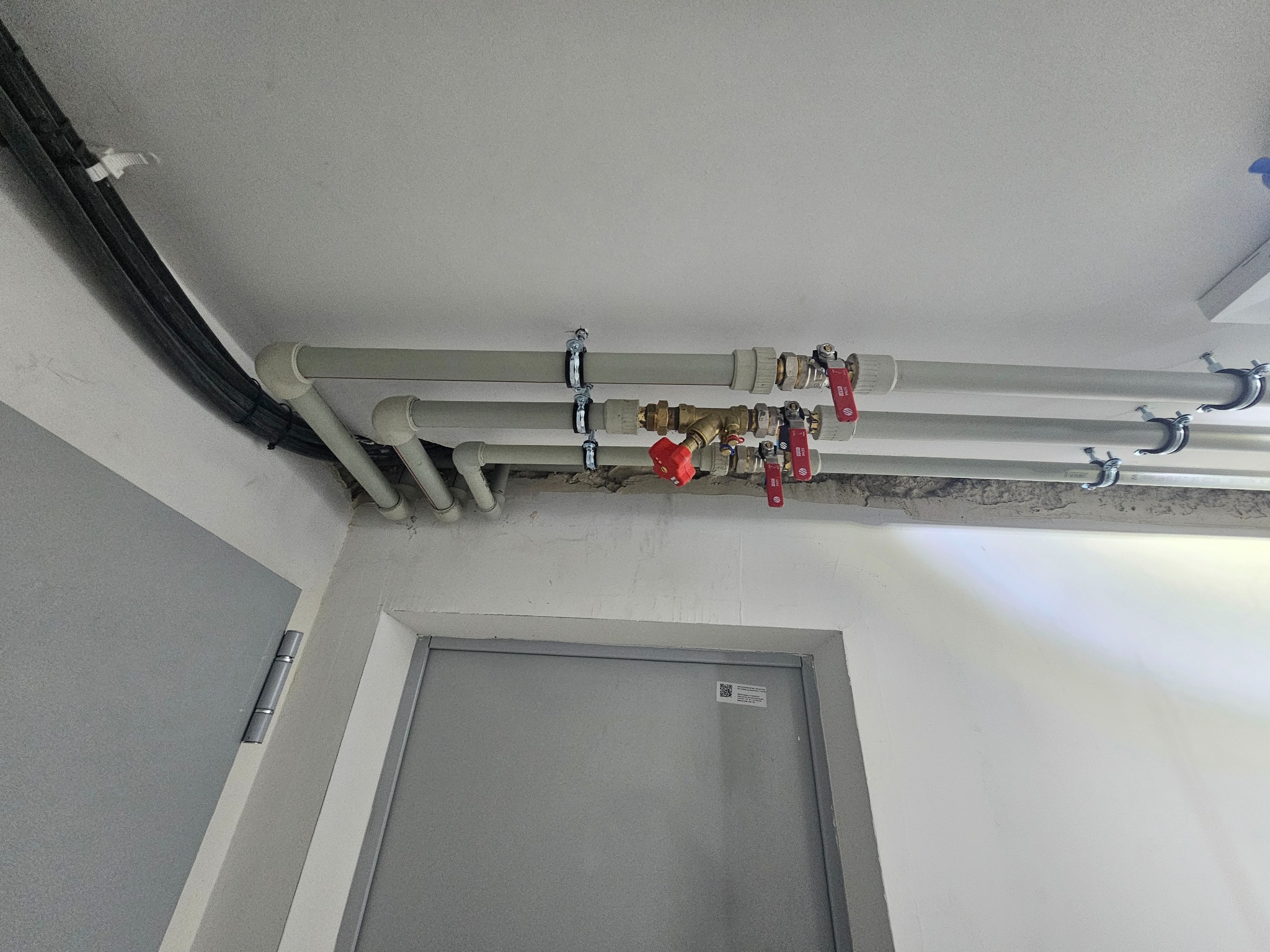 Szare rury instalacji hydraulicznej przymocowane do sufitu, z zaworami z czerwonymi uchwytami, widoczne nad szarymi drzwiami w pomieszczeniu.