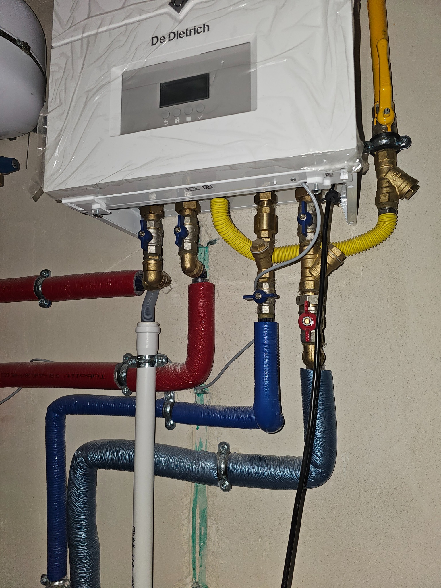 Nowy, biały piec gazowy De Dietrich, jeszcze w folii ochronnej, podłączony do instalacji z rur w kolorach: czerwonym, niebieskim i żółtym. Widoczne zawory i złączki.