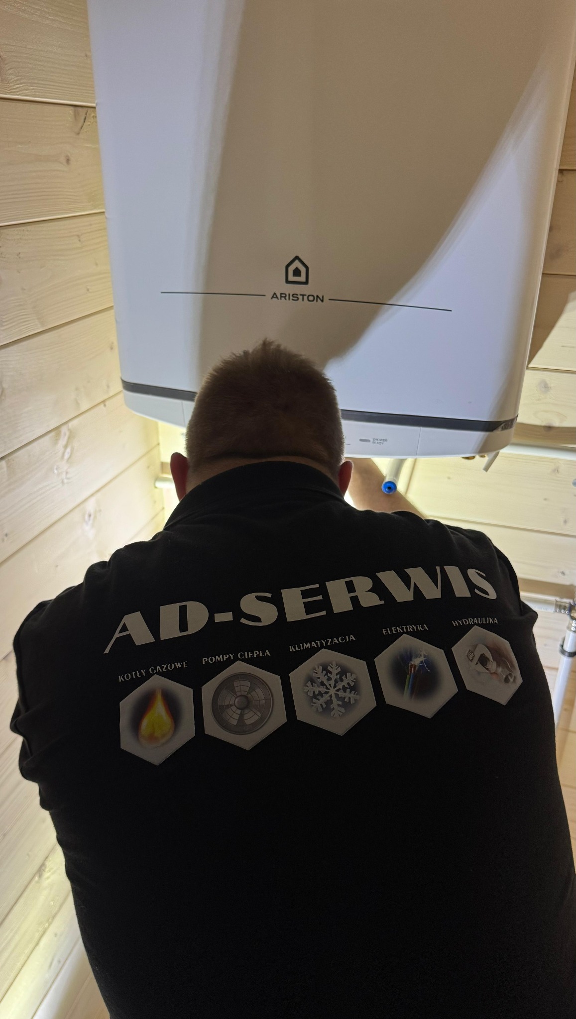 Instalator w czarnej koszulce AD-SERWIS montuje biały piec gazowy Ariston w pomieszczeniu wykończonym boazerią. Widoczne rury instalacyjne.
