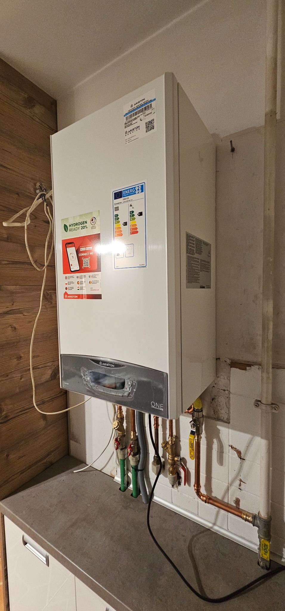 Biały piec gazowy Ariston ONE z widocznymi rurami miedzianymi i zielonymi, zamontowany na ścianie z białymi płytkami, nad blatem kuchennym. Widoczne naklejki informacyjne.