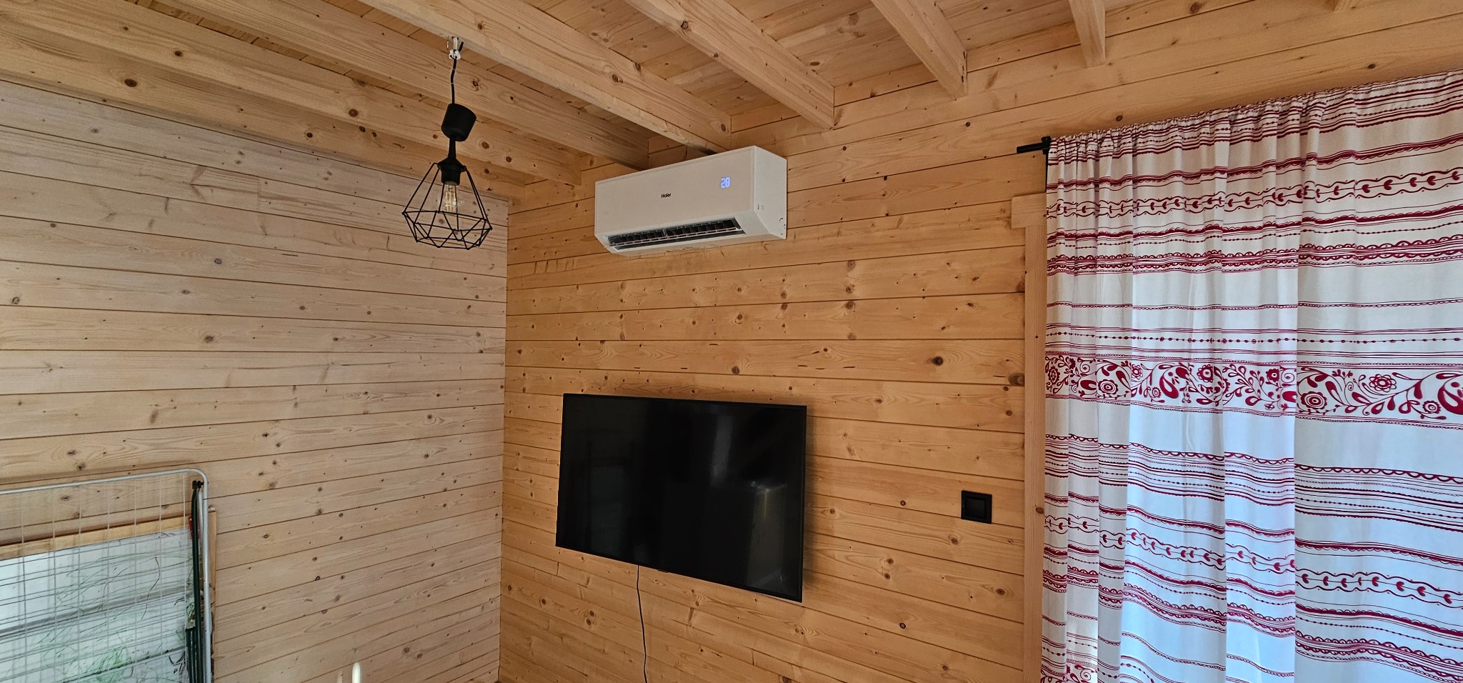 Klimatyzator ścienny Haier zamontowany w drewnianym wnętrzu, z widocznym wyświetlaczem temperatury, lampą wiszącą i zasłoną w czerwone wzory.