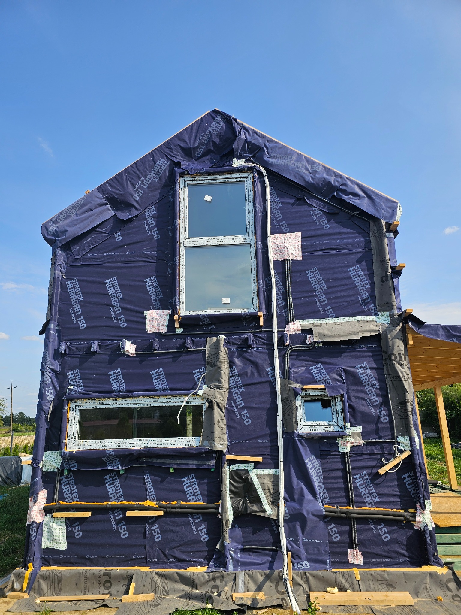 Dom w trakcie budowy, zabezpieczony folią wiatroizolacyjną Stroetex Huragan 150, z widocznymi oknami i instalacją elektryczną na elewacji.