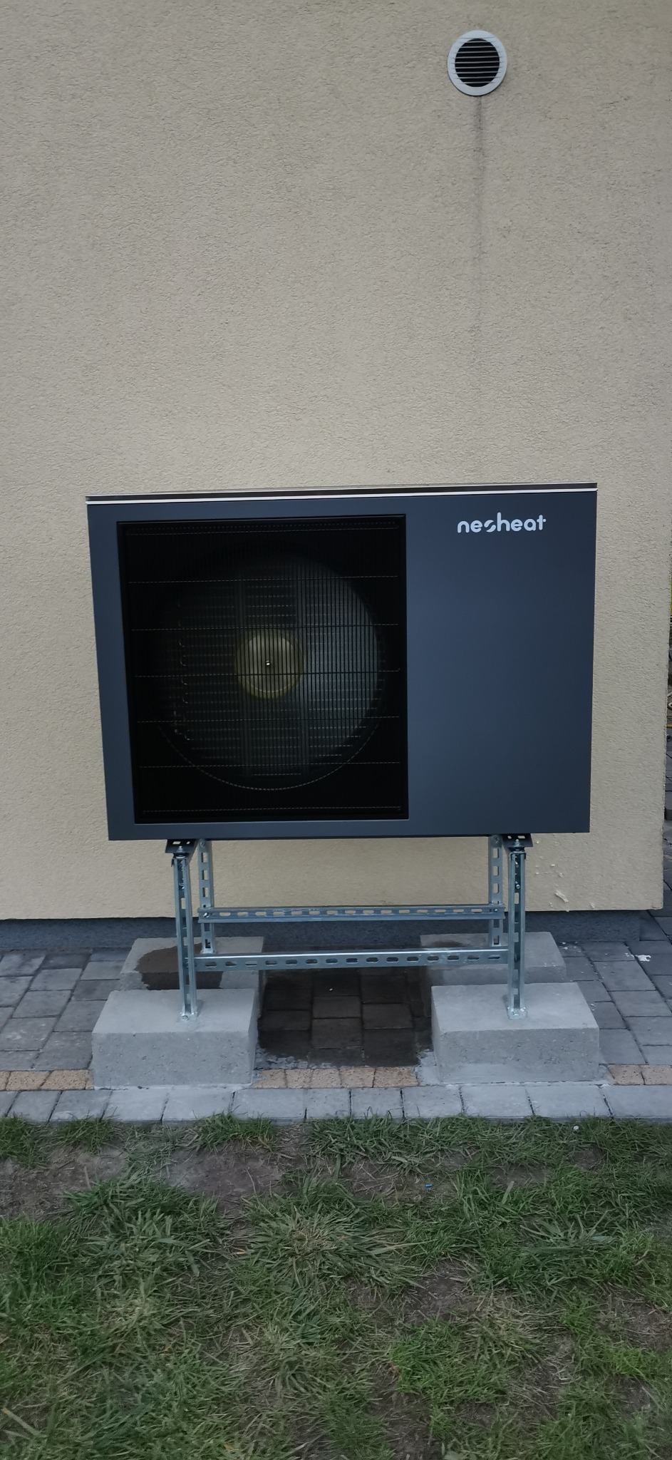 Zewnętrzna jednostka pompy ciepła marki Nesheat, zamontowana na metalowej konstrukcji, na tle elewacji budynku. Widoczny fragment trawnika i kostki brukowej.