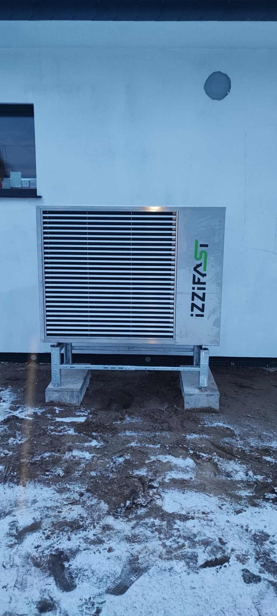 Pompa ciepła IZZIFAST zamontowana na zewnątrz budynku, na betonowych podstawach, z widocznym logo firmy. Śnieg na ziemi.