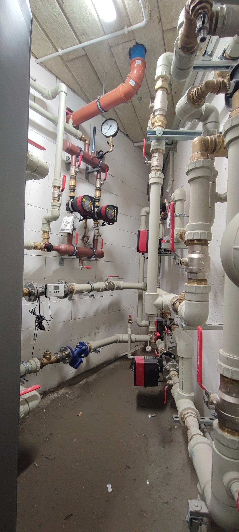 Złożona instalacja hydrauliczna z rur, zaworów, pomp obiegowych i manometru na ścianie z bloczków. Widoczne liczne połączenia i elementy instalacji grzewczej.