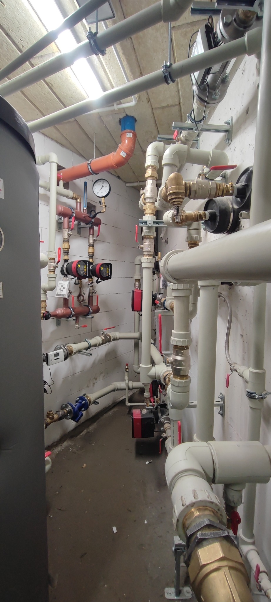 Złożona instalacja hydrauliczna z rurami, zaworami i pompami w kotłowni. Widoczne elementy pomiarowe i zabezpieczające. Różne materiały i kolory rur.