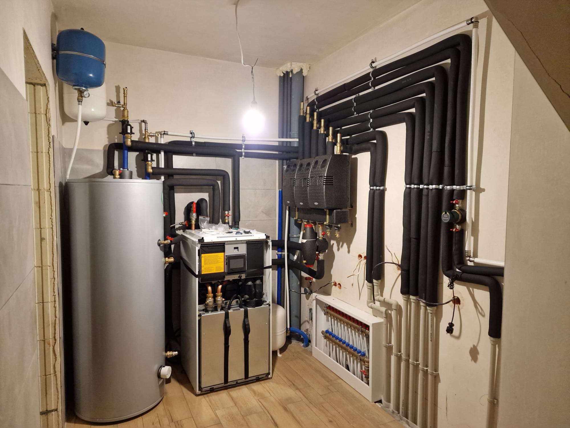 Złożona instalacja grzewcza w pomieszczeniu: piec gazowy, zasobnik CWU, rozdzielacz podłogówki i rury w izolacji. Widoczne liczne przyłącza i zawory.