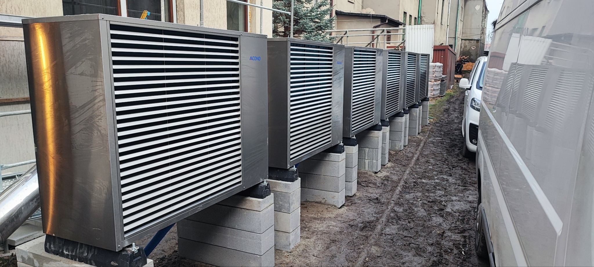 Szereg zainstalowanych pomp ciepła Acond na betonowych podstawach, widoczne z boku na zewnątrz budynku, z pojazdem dostawczym w tle, na błotnistym podłożu.