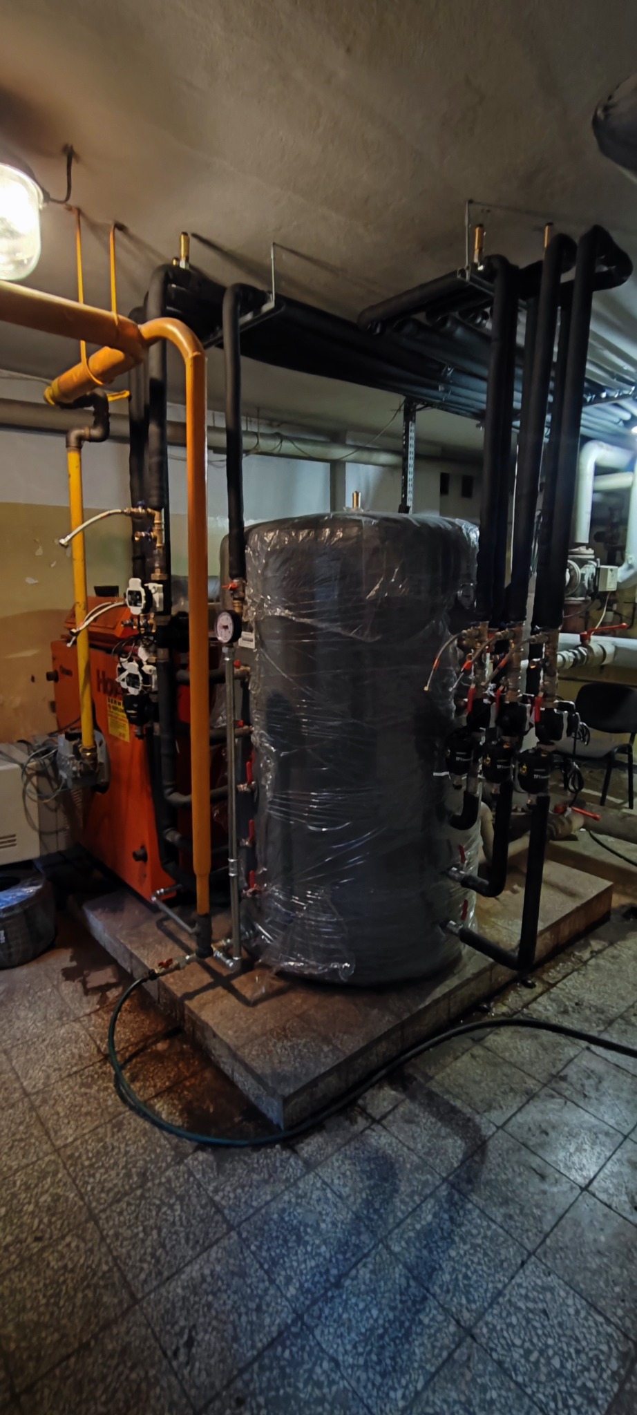 Pomarańczowy piec grzewczy z czarnym zbiornikiem owiniętym folią, połączone rurami w piwnicy z szarą podłogą i białymi ścianami. Widoczne liczne zawory i manometry.