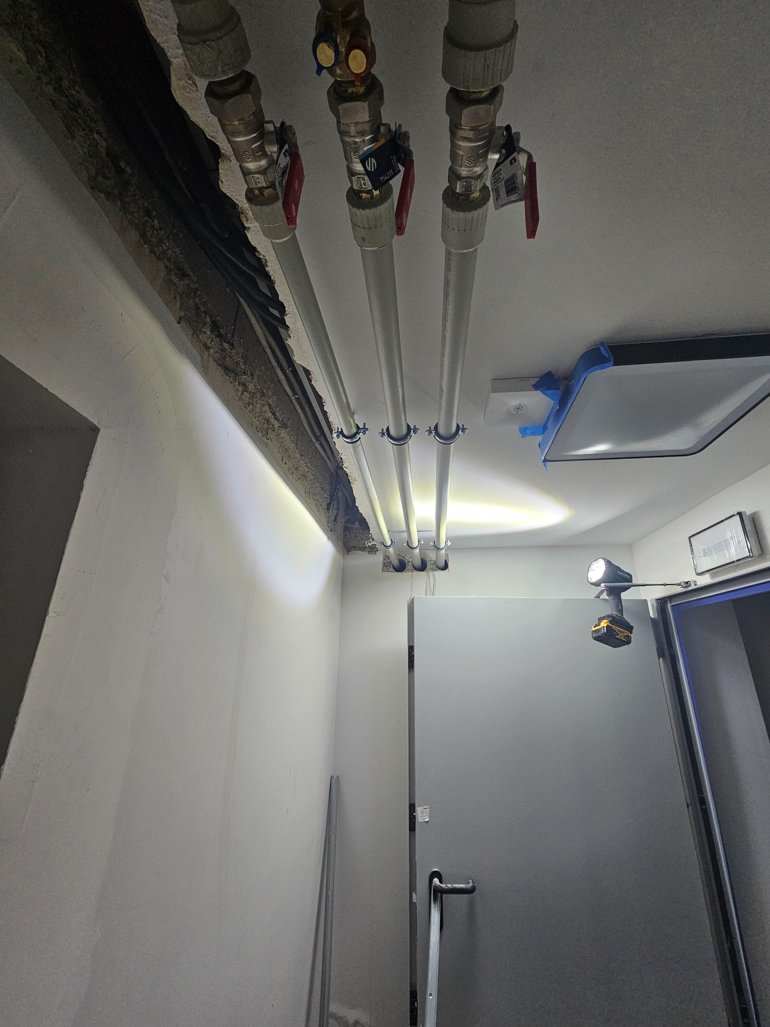 Białe rury z zaworami zamontowane pod sufitem, oświetlone lampą roboczą. Widoczne mocowania i fragment instalacji w ścianie. Prace hydrauliczne w pomieszczeniu.