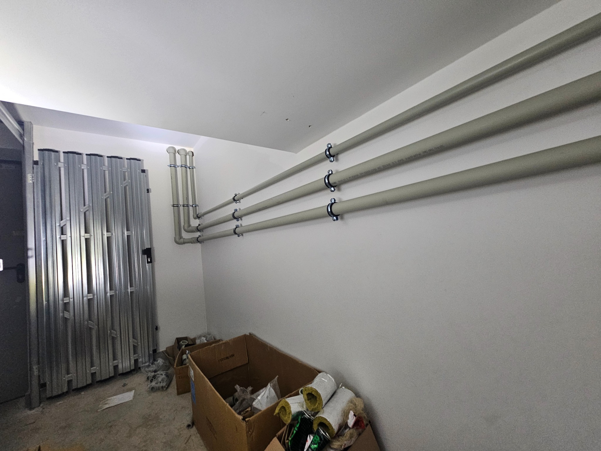 Szare rury instalacyjne przymocowane do ściany za pomocą uchwytów, prowadzone równolegle. Widoczny fragment metalowej bramy i karton z materiałami izolacyjnymi.