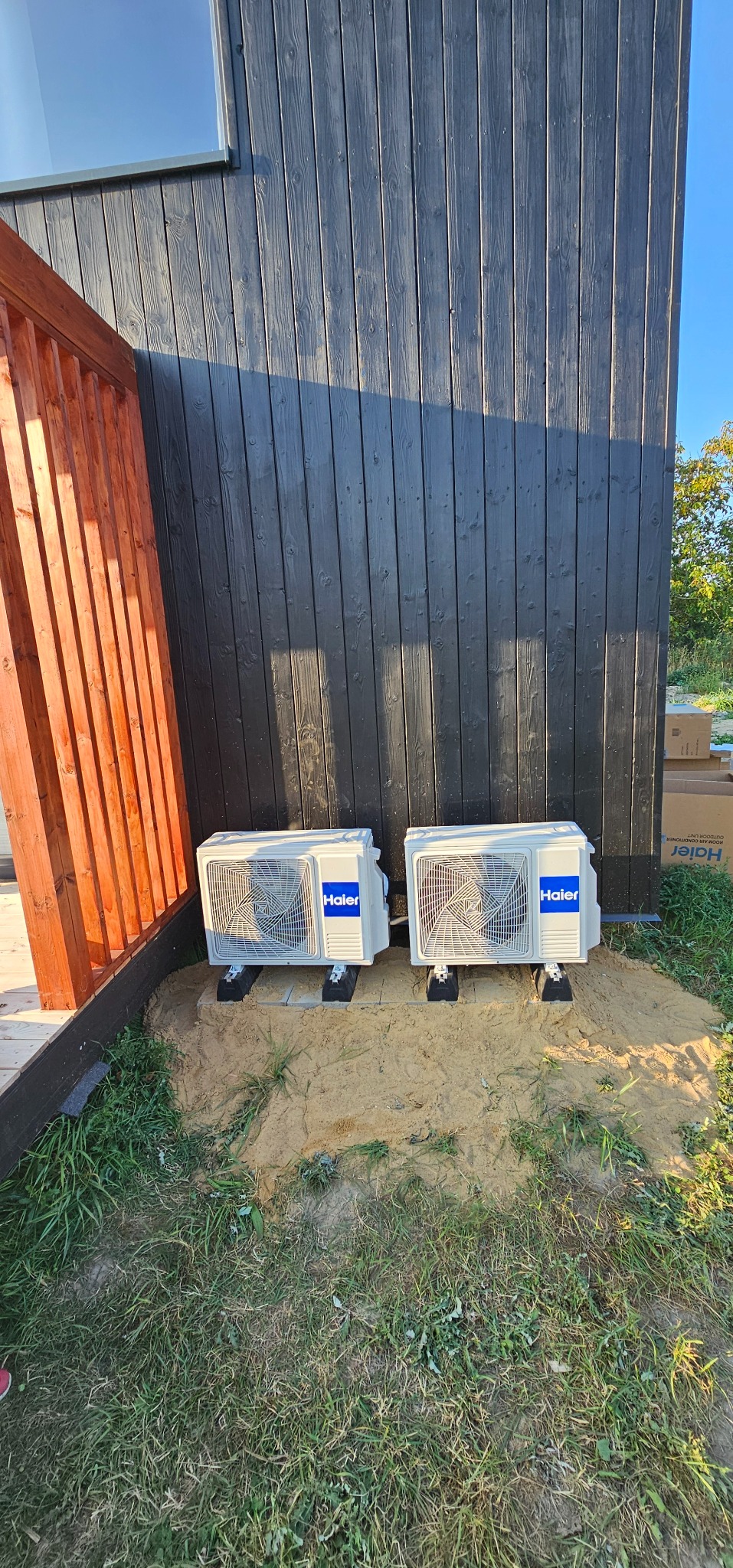 Dwa białe klimatyzatory Haier zamontowane na zewnątrz budynku z ciemnego drewna, na podłożu z piasku i trawy. Widoczne elementy drewnianej konstrukcji.